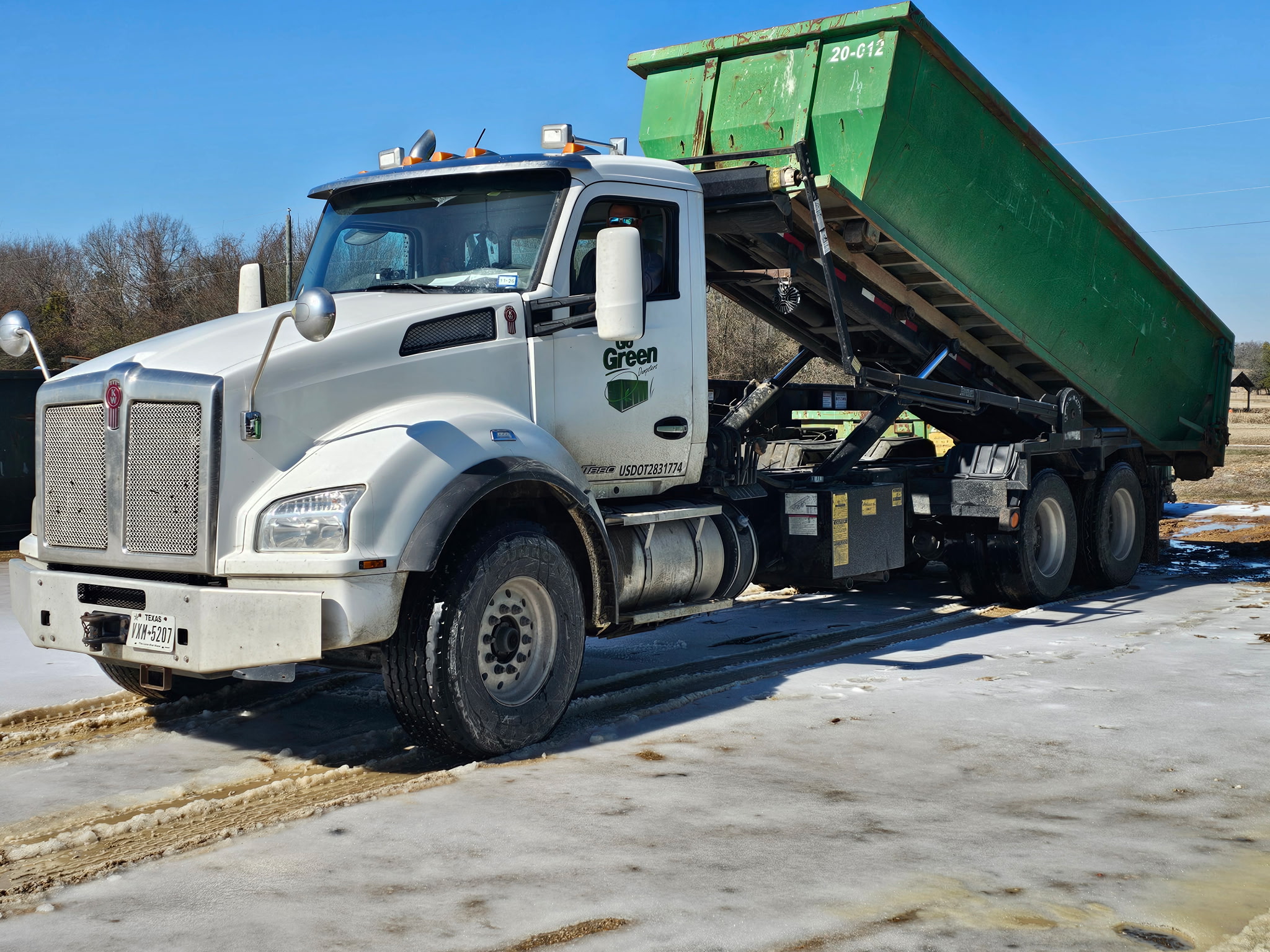 A white dump truck with a raised large green container is parked on a snowy, partially muddy ground under a clear blue sky. Trees and a field are visible in the background.