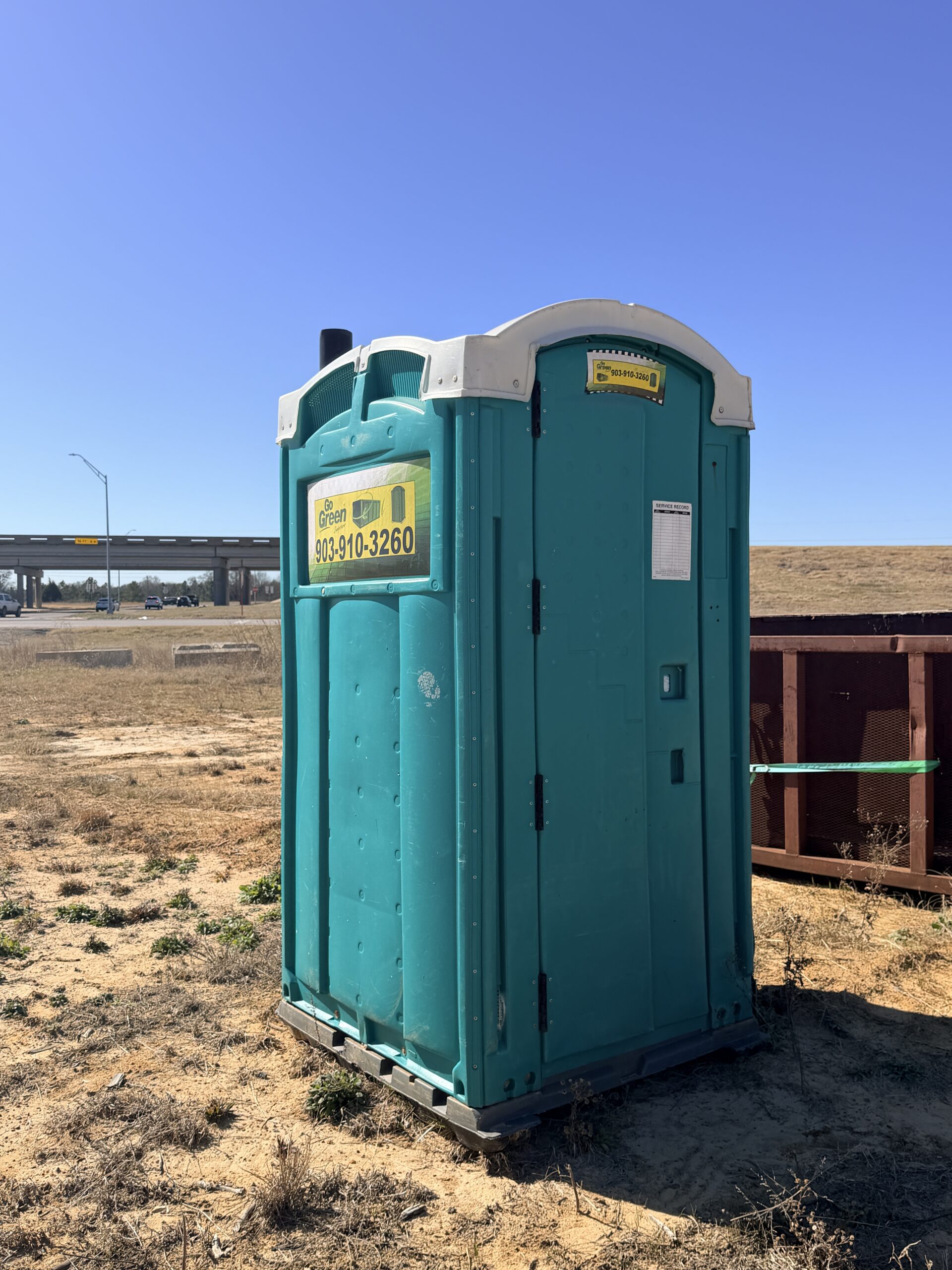 A teal portable toilet stands on dry, sandy ground near a highway overpass and a large dumpster, with sparse grass and clear blue sky in the background.